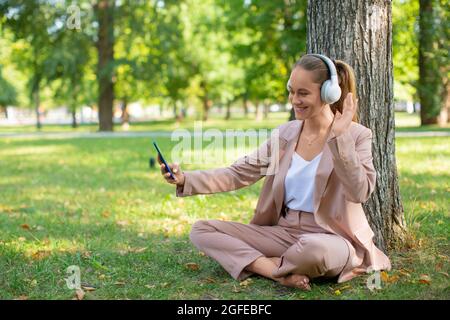 Junge Frau mit Smartphone im Stadtpark während Online-Vido-Anruf. Online-Arbeitskonzept oder Konzept einer entfernten Online-Bildung. Sie schaut sich webc an Stockfoto