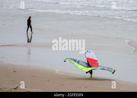 Eine Person, die ein Foil Board trägt; das ist ein Hydrofoil-Surfboard, das normalerweise in Verbindung mit einem Kite zum Antrieb verwendet wird, macht sich seinen Weg entlang des Llangennith Beach, Gower, Swansea, UK, Richtung Meer. Stockfoto