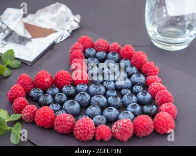 Herzförmiges Liebeszeichen aus frischen Himbeeren und Blaubeeren auf schwarzem Papier. Eine Tafel Schokolade in Folie, Blätter und ein Glas im Hintergrund. Stockfoto