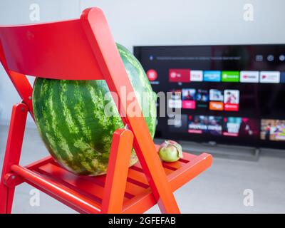 Kleiner winziger roter Apfel mit einem Blatt auf der Oberseite und einer großen Wassermelone, die auf einem roten Holzstuhl sitzt und fernsieht. Stockfoto