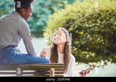 Lächelnde junge Freunde, die sich gegenseitig angusten, während sie auf der Bank sitzen Stockfoto