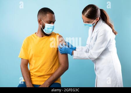 Arzt, der einen afroamerikanischen männlichen Patienten mit Bandhilfe beschimpft, nachdem der Coronavirus-Impfstoff auf blauem Studiohintergrund geschossen wurde Stockfoto