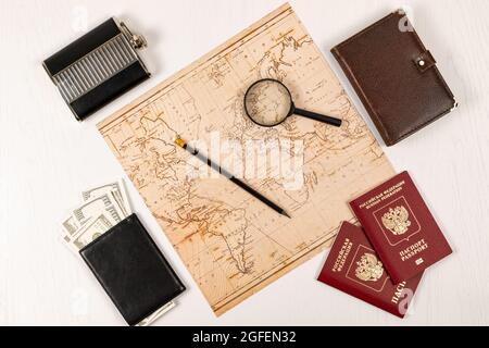 Weltkarte, Bleistift, Lupe, zwei Pässe, Tagebuch, Schwarze Brieftasche mit Geld und eine Flasche auf einem weißen Holztisch. Inschrift - Reisepass Russisch Stockfoto