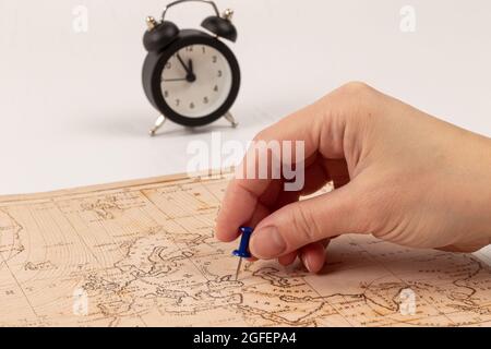 Weibliche Hand zeigt mit einer Nadel den Ort der Reise auf der Weltkarte. Auf einem weißen Holztisch ein schwarzer Wecker. Nahaufnahme. Selektiver Fokus Stockfoto