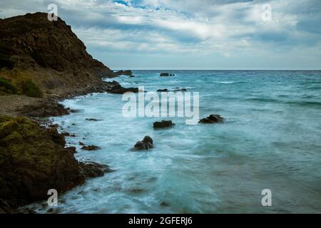 Felsige Strandlandschaft bei Sturm mit bewölktem Himmel. Dramatische, lange Exposition von Meereswellen, die auf Felsen brechen. Stockfoto