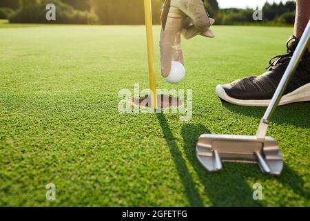 Golfer nimmt Golfball aus Loch in Golfplatz nach erfolgreichem Hit mit Putter, an einem sonnigen Tag im Freien Stockfoto