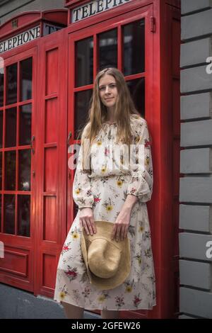 Hübsche kaukasische junge Frau in legerer Kleidung, die in London auf der Straße steht und lächelt. Lange, gerade, blonde Haare. Rote englische Telefonzelle Stockfoto