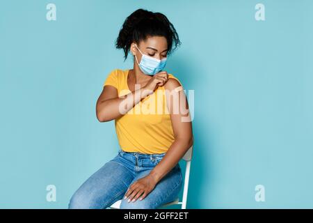 Schwarze Frau in Maske, die Schulter mit Haftverband zeigt und auf blauem Hintergrund gegen Coronavirus geimpft wird Stockfoto
