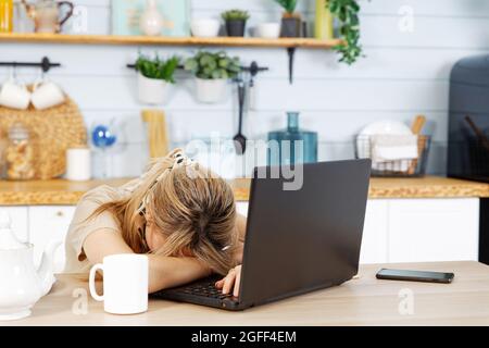 Die junge Frau war müde und schlief am Küchentisch ein, während sie am Laptop arbeitete Stockfoto