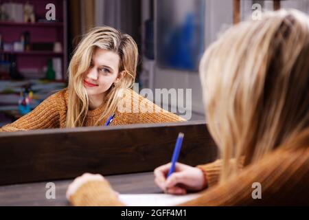 Die junge Frau sitzt an einem Tisch vor einem Spiegel und schreibt etwas mit einem Stift in ein Notizbuch. Geringer Fokus. Stockfoto