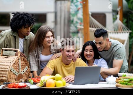 Eine Gruppe von Millenniumsfreunden mit mehreren Rassen, die im Herbst einen Laptop auf dem Campingausflug aufs Land benutzen, online chatten und Spaß haben Stockfoto