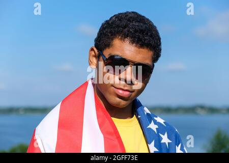 Mann, der die Kamera anschaut und stolz die amerikanische Flagge auf seinen Schultern hält Stockfoto