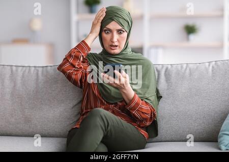Schockierte Muslimische Frau, Die Zu Hause Smartphone Hält Und Den Kopf Berührt Stockfoto