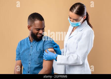 Der junge Arzt in einer Gesichtsmaske legt nach dem Schuss des Covid-Impfstoffs auf beigen Hintergrund ein Pfändelhilfsmittel auf den Arm des schwarzen männlichen Patienten Stockfoto