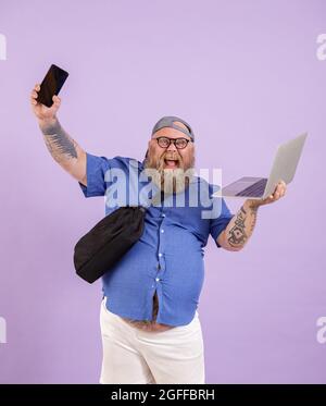 Positiver Plus-Size-Mann mit Laptop und Tasche zeigt Telefon auf violettem Hintergrund im Studio Stockfoto