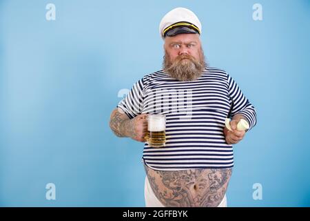 Lustige übergewichtige Seemann hält rauchende Pfeife und Bier Grimacing auf hellblauem Hintergrund Stockfoto