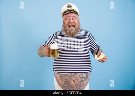 Fröhliche Plus-Größe Person im Matrosenanzug hält Rohr und Bier auf hellblauem Hintergrund Stockfoto