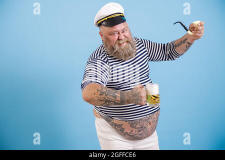 Grinsender Segler in Übergröße hält rauchende Pfeife und leckeres Bier auf hellblauem Hintergrund Stockfoto