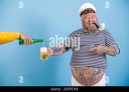 Fröhlicher Seemann mit Übergewicht hält Becher, während Frau Bier auf hellblauen Hintergrund gießt Stockfoto