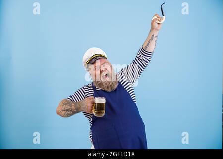 Aufgeregt übergewichtiger Mann Seemann hält Bier und hebt Rohr auf hellblauem Hintergrund Stockfoto