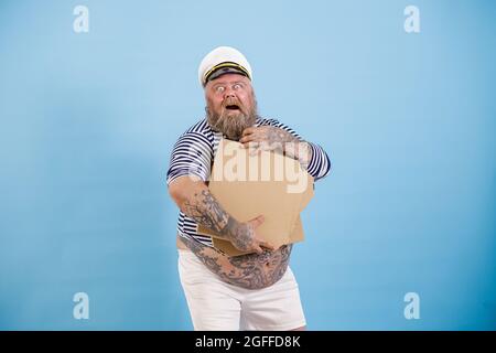 Glücklicher bärtiger übergewichtiger Mann Seemann hält leere Kartons mit Pizza, die auf hellblauem Hintergrund posieren Stockfoto