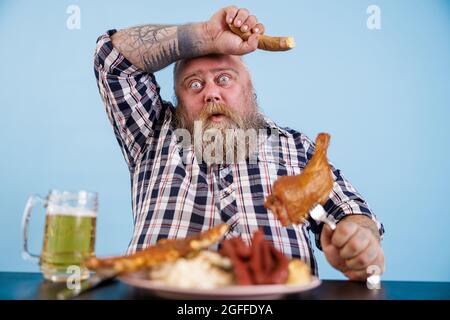 Müde übergewichtiger Mann wischt die Augenbrauen, die unter übermäßigem Essen am Tisch mit fetten Speisen im Studio leiden Stockfoto