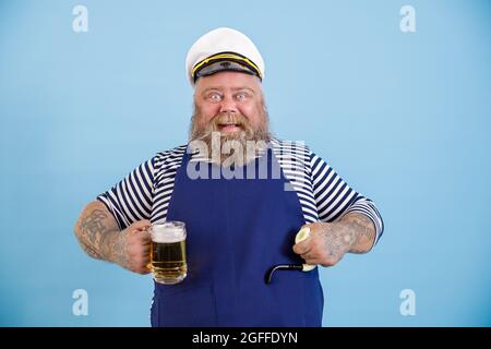 Lustige dicke Person im Matrosenkostüm hält rauchende Pfeife und Bier auf hellblauem Hintergrund Stockfoto