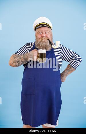 Positiv fetter Mann im Matrosenanzug mit Schürze hält Craft-Bier auf hellblauem Hintergrund Stockfoto