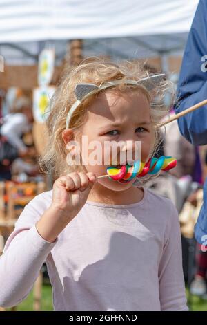 Mädchen essen bunte Süßigkeiten. Ein fünfjähriges Mädchen in rosa Kleidung isst eine bunte Süßigkeit auf einem Stock. Sonniger Sommertag Stockfoto