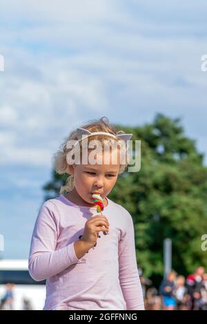 Mädchen essen bunte Süßigkeiten. Ein fünfjähriges Mädchen in rosa Kleidung isst eine bunte Süßigkeit auf einem Stock. Sonniger Sommertag Stockfoto