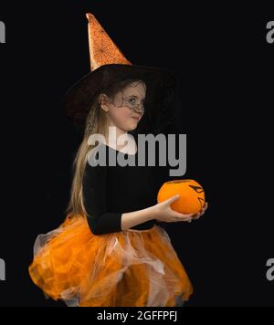 Ein lustiges Mädchen mit einem Jack Kürbis und in einem Hexenkostüm für Halloween-Feier. Orangefarbener Hut und Rock, schwarze Bluse, Spinnennetz-Schleier, schwarzer Hintergrou Stockfoto