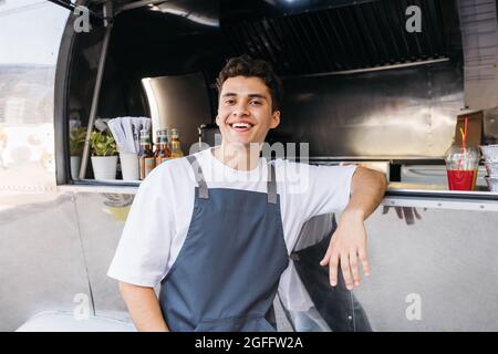 Porträt eines glücklichen Besitzers eines Food-Trucks. Typ aus dem Nahen Osten im Vorfeld, der sich auf einen Food Truck lehnte und die Kamera ansah. Stockfoto