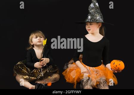 Zwei Schwestern verkleidet als Hexen. Eine schwarze Bluse, ein Hut mit Spinnweben, ein oranger Rock. Das ältere Mädchen hält einen Kürbiskübel für Süßigkeiten, den jüngeren Stockfoto