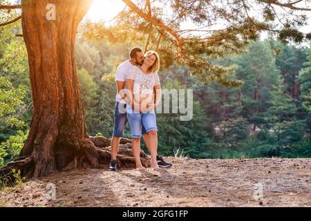 Junges erwartungsdiges Paar, das bei Sonnenuntergang die Hände am Bauch unter dem Baum hält Stockfoto