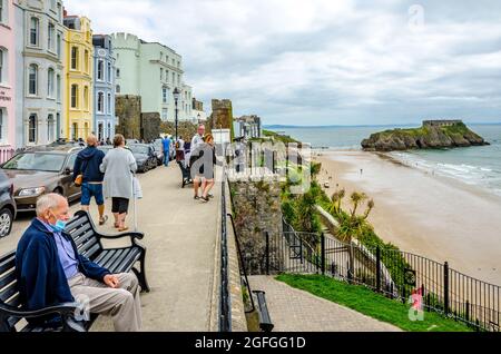 Ein Mann sitzt auf der Bank auf der Esplanade in Tenby, Reading mit dem South Beach und St. Catherine's Island in der Ferne. Stockfoto