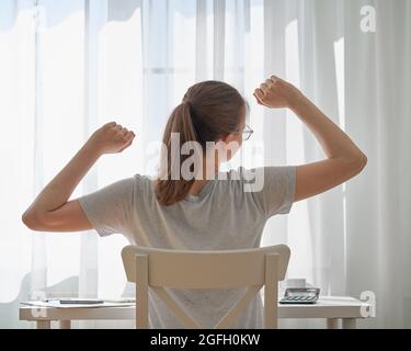 Junge Frau, die sich beim Sitzen am Schreibtisch zu Hause dehnt. Remote-Arbeit Herausforderungen und Unbehagen Stockfoto