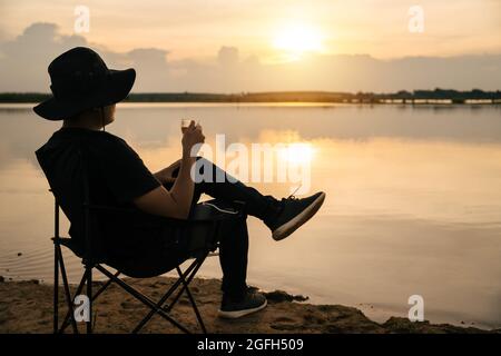 Asiatischer junger Mann, der in einem Touristenstuhl sitzt und eine Tasse mit Tee oder Kaffee hält, den Blick auf die Landschaft des Flusses bei Sonnenuntergang genießt. Tra Stockfoto