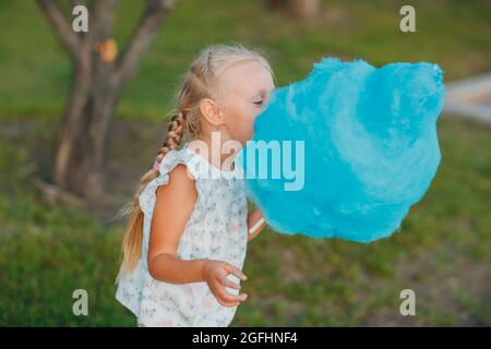 Kleines blondes Mädchen, das im Park blaue Zuckerwatte isst Stockfoto