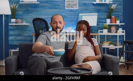 POV der interracial Familie Blick auf Kamera auf der Couch sitzen. Gemischtes Rennpaar erwartet Kind beim Fernsehen. Kaukasischer Mann bringt Glas Wasser für schwarze Schwangere Stockfoto
