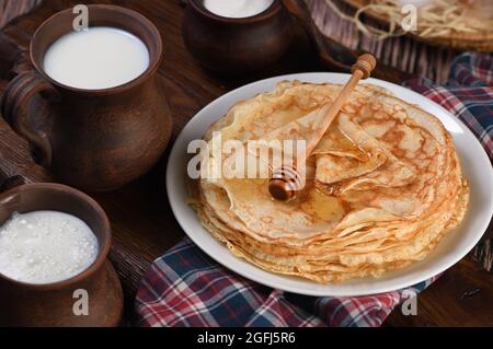 Hausgemachte dünne Pfannkuchen mit Honig, gestapelt in einem Stapel, auf einem Holztisch mit einem Becher Milch, einem Topf mit saurer Rahm und Eiern in einem Korb. Traditionell Slawisch Stockfoto