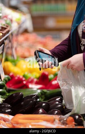 Super U Supermarkt: Frau, Kommissionierer / Packer Auswahl von Produkten in der Obst- und Gemüseabteilung eines Supermarkts mit einem mobilen Gerät zu c Stockfoto