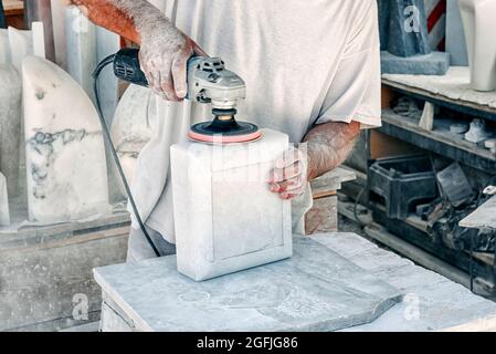 Der Mann, der während des Produktionsprozesses einen Marmorblock in einer Werkstatt aus nächster Nähe an seinen Händen und dem Elektrowerkzeug mit einem Schleifgerät aus dem Orbitalschleifer glättet Stockfoto