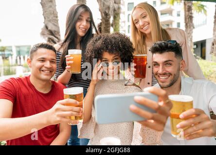 Eine Gruppe von Freunden, die für ein Selfie posieren und dabei mit einem fröhlichen Lächeln ihre Gläser auf die Kamera auf dem Handy heben Stockfoto