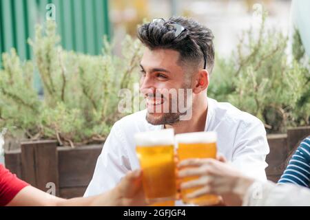 Trendiger lächelnder Mann, der mit seinen Freunden in einem Pub im Freien feiert, während sie ihre Gläser kaltes Bier in einem Toast mit Fokus auf die Getränke zu seinem Freund heben Stockfoto