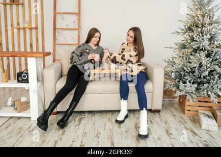 Zwei junge Frauen unterhalten sich und trinken Tee auf dem gemütlichen Sofa. Weihnachtsdekoration auf dem Hintergrund. Stockfoto