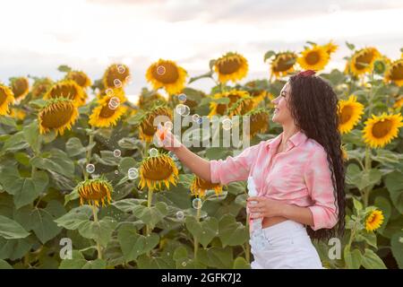 Nette Brünette Frau bläst Seifenblasen. Schönes Feld mit Sonnenblumen auf dem Hintergrund. Stockfoto