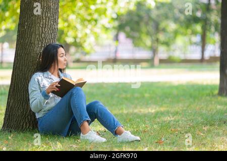 Asiatische Frau mit einem Buch im Park. Sie sitzt auf dem Gras im Park und schaut nebeneinander. Das Konzept der Ruhe von Gadgets, Smartphones und der Inte Stockfoto