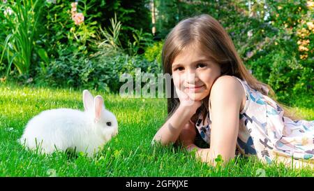 Ein schönes Mädchen liegt auf dem Rasen und spielt mit einem kleinen Hasen. Dekoratives Zwergkaninchen, das Gras auf dem Rasen im Freien frisst. Freundschaft zwischen Kindern und Haustieren Stockfoto