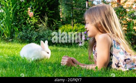 Ein dekorativer Zwergkaninchen frisst Gras auf dem Rasen im Garten. Das Mädchen liegt auf dem Rasen und spielt mit einem niedlichen weißen Hasen. Nagetier als Haustier. Chil Stockfoto