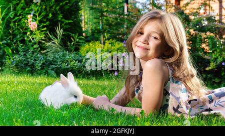 Das Mädchen lächelt scharf. Im Sommer spielt ein Kind mit einem weißen flauschigen Hasen im Garten auf einer grünen Wiese. Dekorativer Zwergkaninchen, der Gras auf dem l frisst Stockfoto